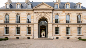 Façade en pierre d’un hôtel particulier français avec porte cochère et cour d’honneur pavée, éclairée par une lumière naturelle.