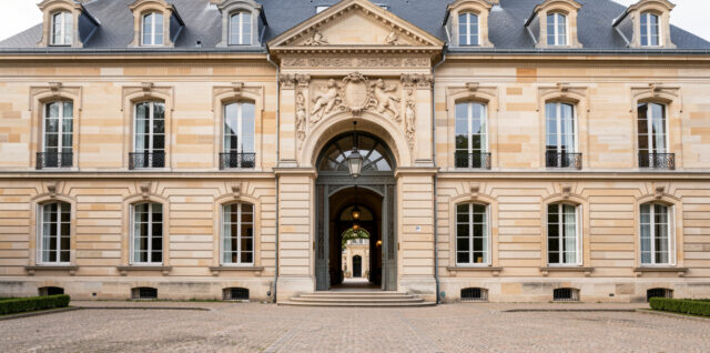 Façade en pierre d’un hôtel particulier français avec porte cochère et cour d’honneur pavée, éclairée par une lumière naturelle.